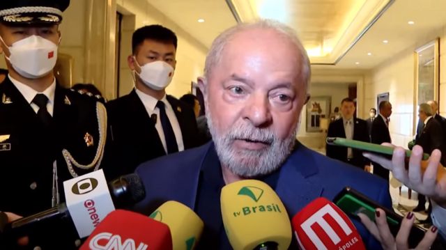 Lula dando entrevista em saguão de hotel, observado por dois guardas chineses e rodeado por microfones 