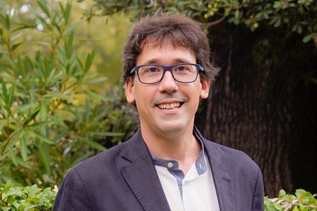Retrato do economista português Pedro Maia Gomes. Ele usa uma camisa branca e um terno cinza escuro em um jardim.