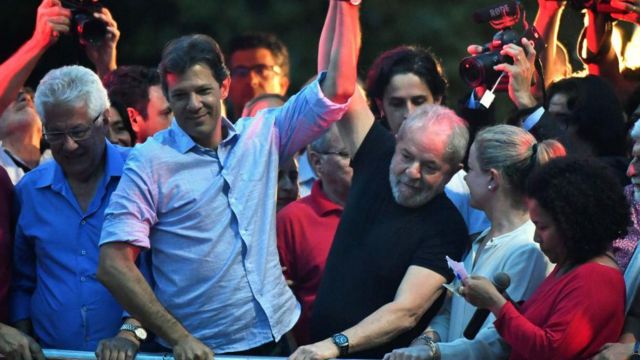 Haddad e Lula levantando os braços juntos, rodeados por outras pessoas em evento