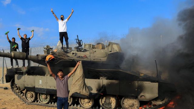 Palestinos comemoram enquanto um veículo militar israelense pega fogo depois de ter sido atingido por homens armados que se infiltraram em áreas do sul de Israel, no lado israelense da fronteira 