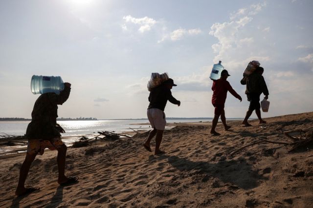 Sombras de quatro pessoas carregando galões d'água em terra seca na margem de rio