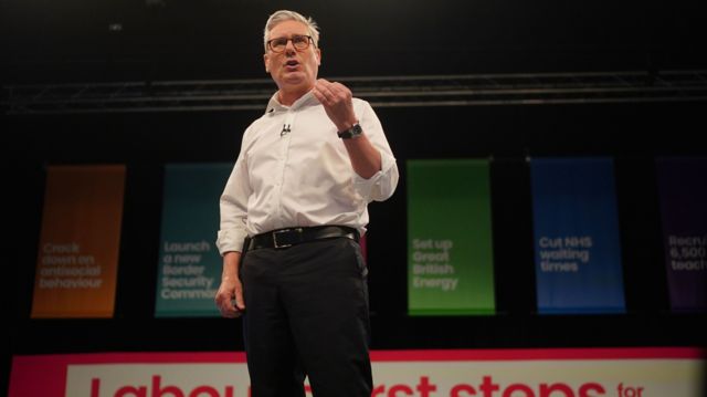 Sir Keir Starmer doing a speech stood in front of a backdrop showing Labour's six pledges