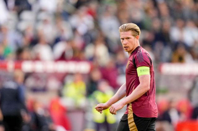 Belgium captain Kevin De Bruyne in action for his country in a friendly.  He has a focused expression on his face.  He is wearing a bright captains armband.  He has a burgundy Belgium jersey on with gold trimming
