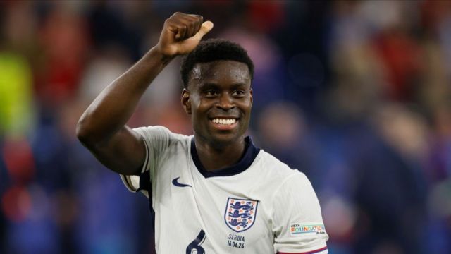 Marc Guehi celebrating after England win against Serbia at Euro 2024.  He is wearing a white england top.  His right hand is raised in celebration and he has a smile on his face.