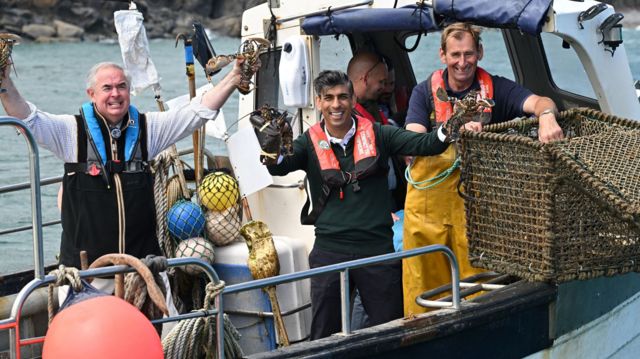 Prime Minister Rishi Sunak smiles as he holds a lobster in each hand while on a boat in the harbour at Clovelly.