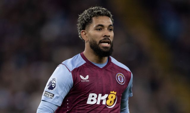 Douglas Luiz, with beared and partially dyed short blonde hair, looks at something out of the picture wearing Aston Villa's claret and blue home shirt