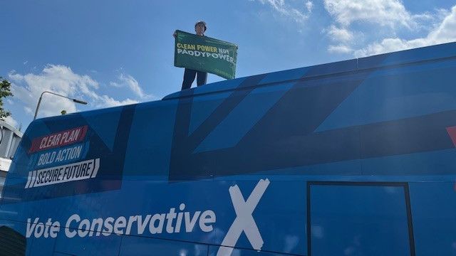 Greenpeace activist holding sign which reads: "Clean power not Paddypower", standing on top of Tory party-branded coach