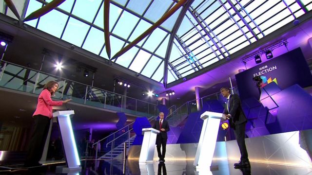 A wide-shot showing host Mishal Husain speaking to Rishi Sunak, with Keir Starmer looking over at Sunak.