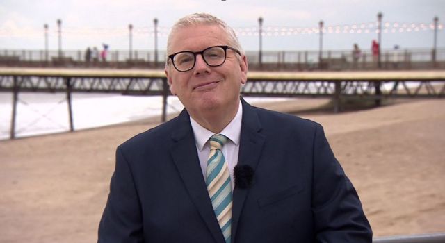 Tim Iredale smiling with a suit and tie and glasses on on a beach with a microphone on his jacket