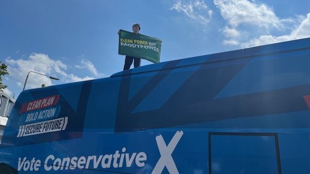 Greenpeace protester on Tory Battle Bus with sign reading: "Clean Power not Paddy Power"
