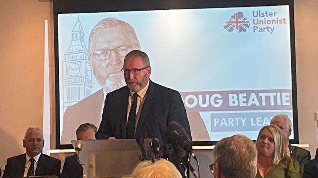 Ulster Unionist Party leader Doug Beattie standing at a podium at an event, with supporters seated around him.