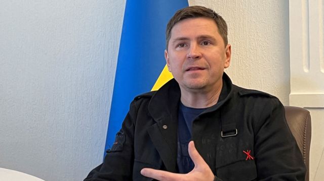 Mykhailo Podolyak sat in a chair talking with a Ukraine flag behind him
