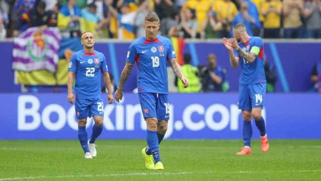 Three Slovakia players on the pitch during Euro 2024.  The are wearing a blue jersey with red trimming and white numbers.  They look annoyed after losing to Ukraine
