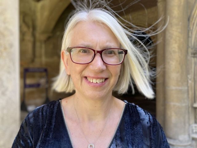 Clare Smalley with glasses and a blue dress and necklace on smiling in the grounds of Lincoln Cathedral