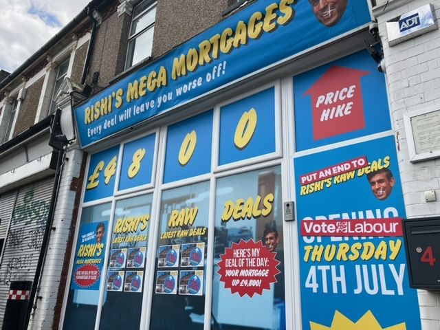 A shop called Rishi’s Mega Mortgages plastered with blue posters. Below the shop sign it says Every deal will leave you worse off.