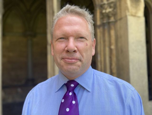Karl McCartney smiling with a blue shirt and purple tie with white dots on in the grounds of Lincoln Cathedral