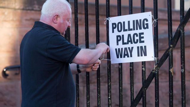 A man in a black t shirt hanging a sign reading polling place way in on some black railings.
