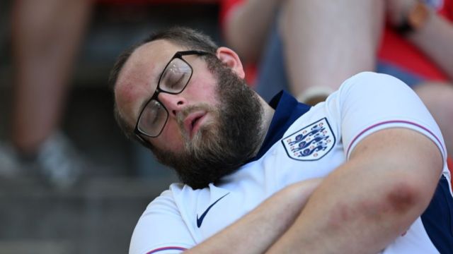 England fan looks like he is sleeping during a Euro 2024 match.  He has his head tilted to the side with his eyes closed.  He is wearing a white England top