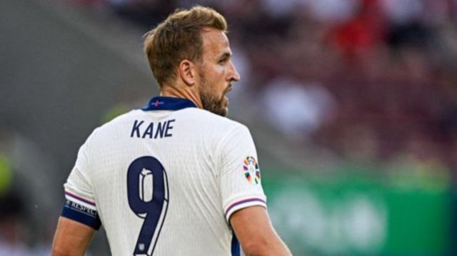 Harry Kane stands with his back towards the camera during a Euro 2024 game for England.  He is wearing a white jersey with a blue number 9 on his back.