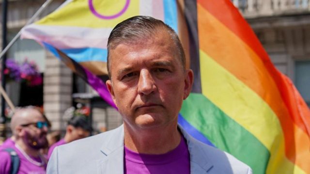 Craig Jones looking at the camera, wearing a purple shirt, blue blazer and his military badges with a Pride flag behind him and a group of marchers on the road as part of Pride in London