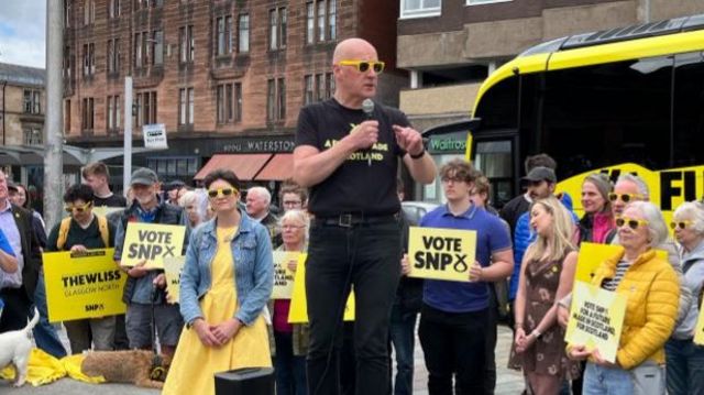 SNP leader John Swinney speaking into a microphone with a group of people behind him. There is a yellow campaign bus behind.