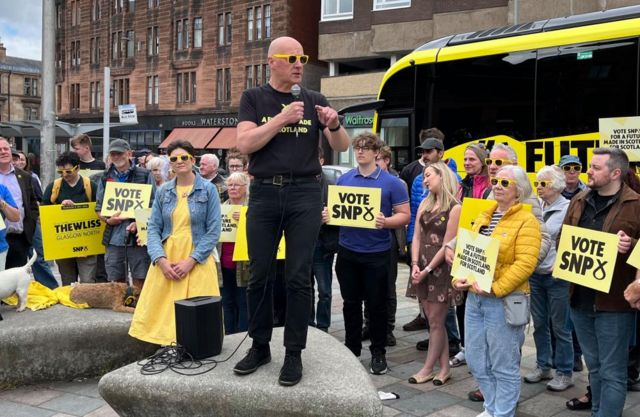 SNP leader John Swinney standing on a concrete block holding a microphone speaking to a crowd, with supporters standing behind him holding yellow Vote SNP signs