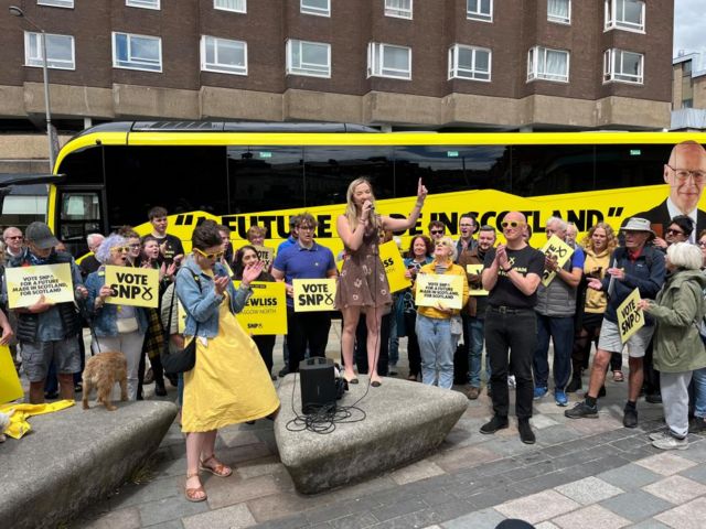 Singer Iona Fyfe speaking in front of a group of people and the SNP campaign bus