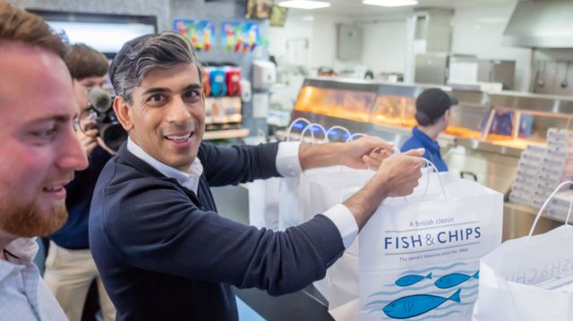 Rishi Sunak holds two bags labelled fish and chips while smiling at the camera