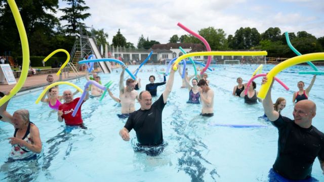 Sir Ed Davey in a swimming pool waving a yellow flotation device during a water aerobics class in Cheltenham