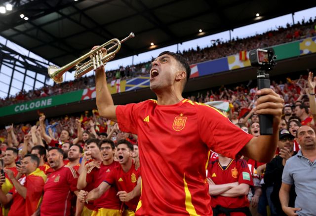 A Spain fan holds up a trumpet and a camera