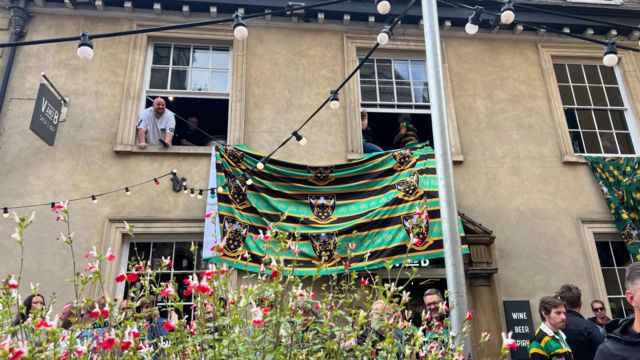 A man looking out of a window with a Northampton Saints flag on display