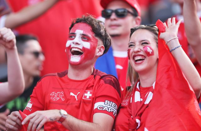 Two Switzerland fans are all smiles after their sides last 16 victory over Italy during Euro 2024.  They both have their faces painted red and are wearing a red Swiss football top.