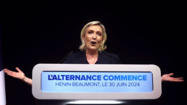 Marine Le Pen reads speaks from a podium featuring phrase 'L'alternance commence'