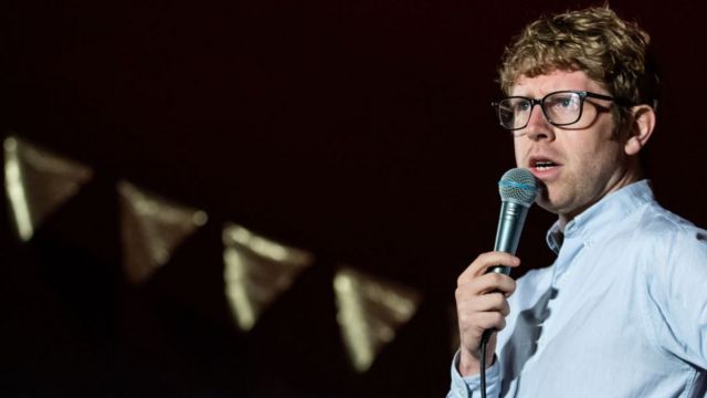 Comedian Josh Widicombe standing with a microphone towards his mouth
