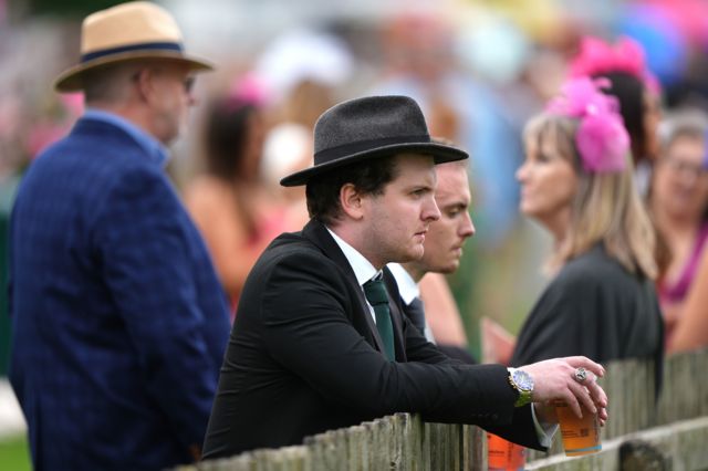 A general view of a minute's silence being observed at Newmarket Racecourse. There is a racegoer in the foreground holding a drink and dressed in a black suit