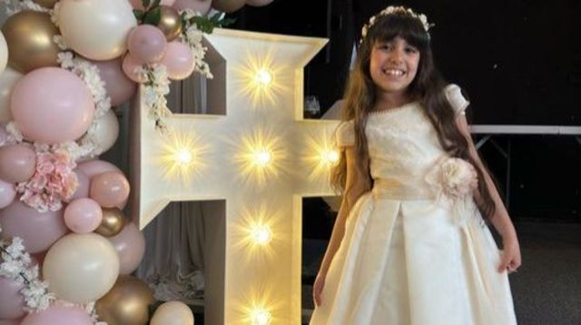 Alice Aguiar wears a white dress and stands next to a cross