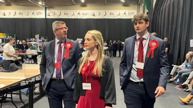 Rosie Wrighting with long blond hair and red dress flanked by two men