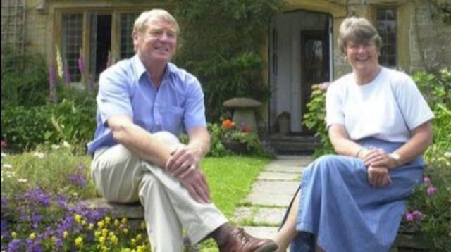 Paddy Ashdown and Jane Ashdown sat on a wall outside a Cotswold stone house with a garden full of flowers.
