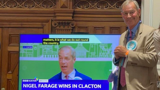 Rupert Lowe standing in front of a TV screen showing Nigel Farage wins in Clacton