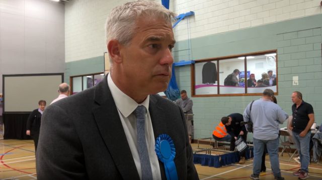 Stephen Barclay looking concerned, dressed in a suit and blue tie, with a blue rosette attached to his lapel.