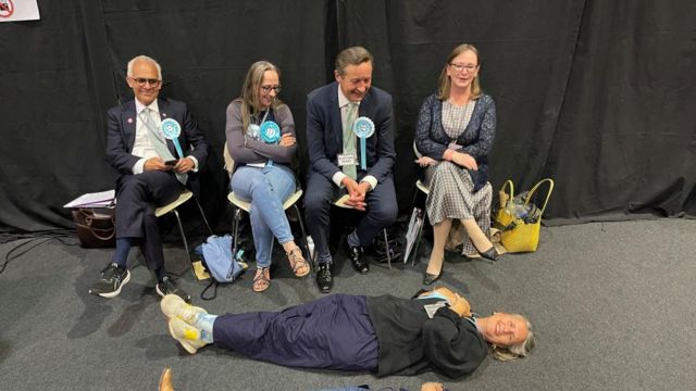 Four seated Reform UK supporters look at a woman lying on the floor
