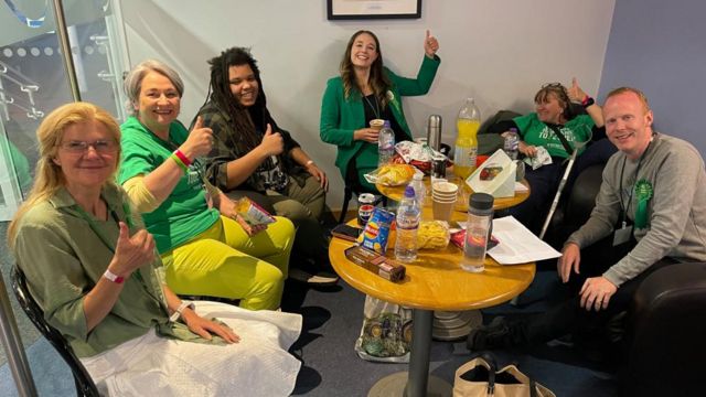 Seven people, some wearing green, sit round a table with water bottles, crisps and biscuits