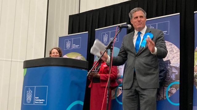 Anthony Browne, with his hands raised, speaks into a microphone on a platform and behind a podium. He is wearing a grey suit with a blue tie and blue rosette pinned to his lapel.