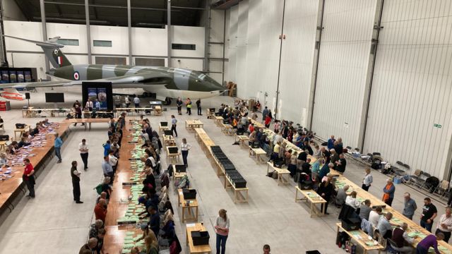 Rows of tables with ballot counters going through ballot papers in the hangar, with a green and blue plane in the background.