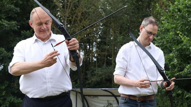 Liberal Democrat leader Sir Ed Davey and Ian Sollom try archery, outside and with woodland behind them.