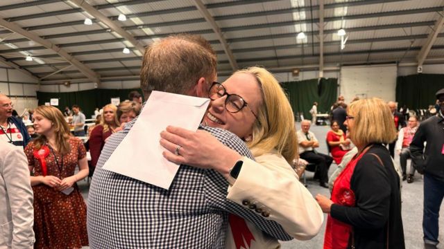 Lucy Rigby with long blond hair and Labour rosette hugs a man at a count