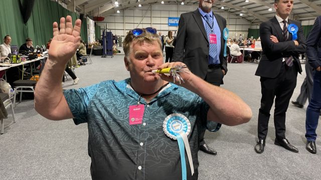 Man with short hair and blue shirt with Reform rosette blows a party whistle