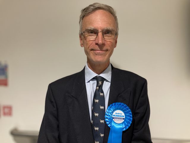 Andrew Murrison smiling while wearing a blue rosette with his name on it at the count