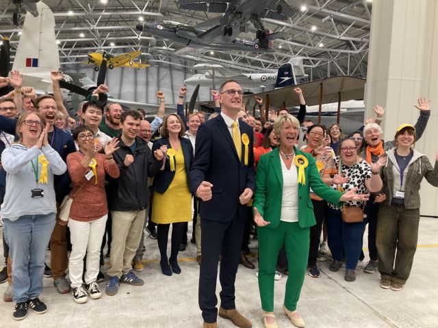 Pippa Heylings in a green suit cheering, standing alongside a male in a suit and a number of cheering supporters behind her.