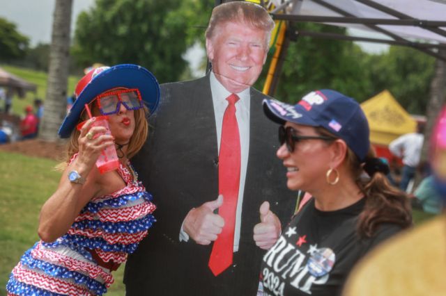 A woman holding a cup leans against a cardboard cutout of Donald Trump